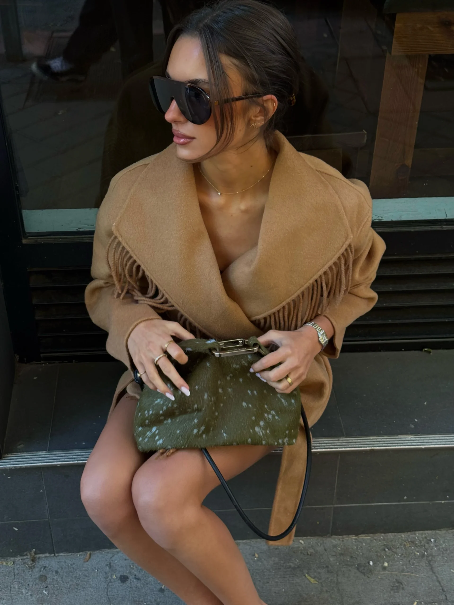 Woman in camel fringe wrap coat with oversized sunglasses and olive-green handbag on her lap.