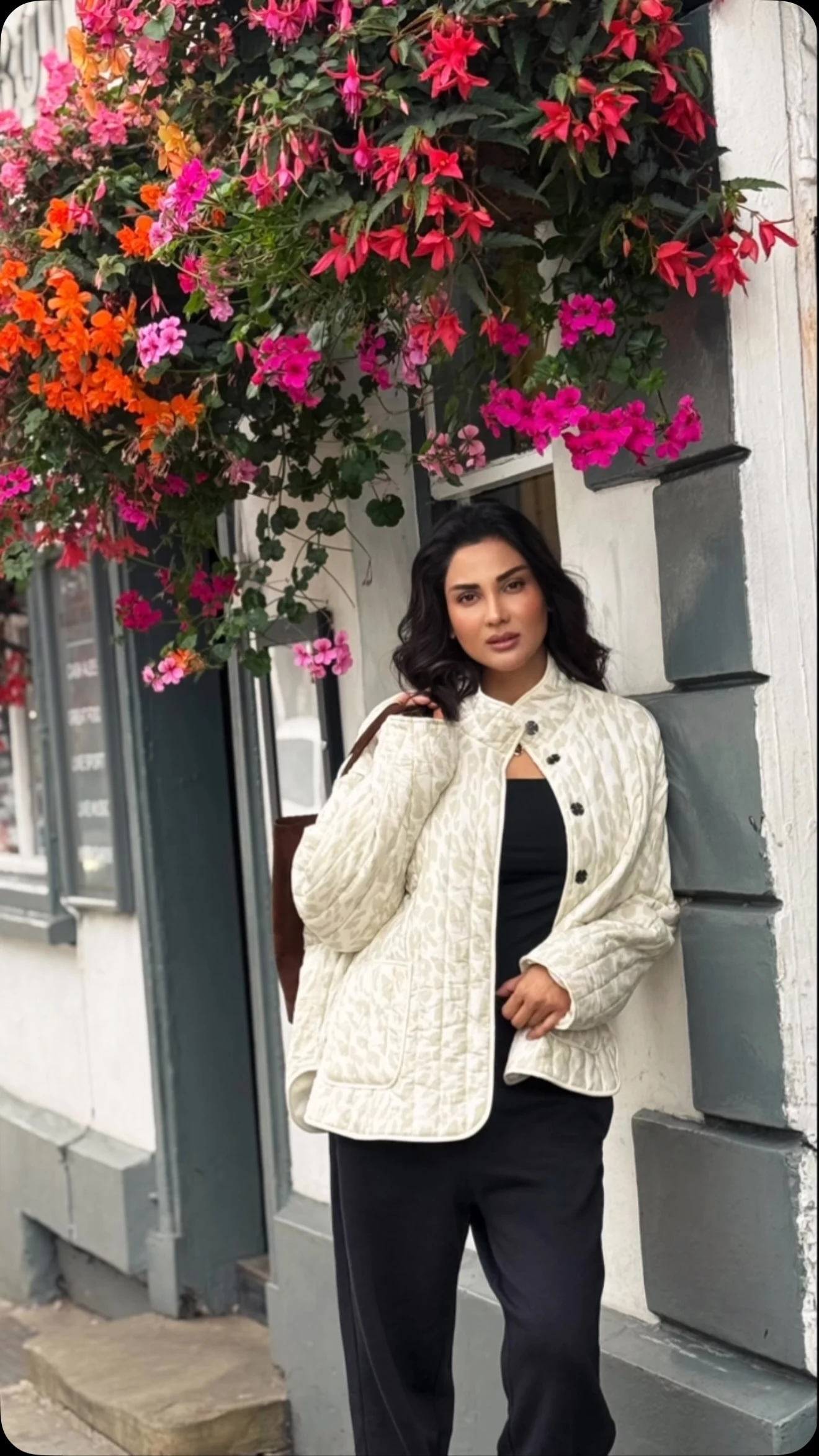 Woman in open cream quilted jacket over black top and trousers beside a flower-covered building.