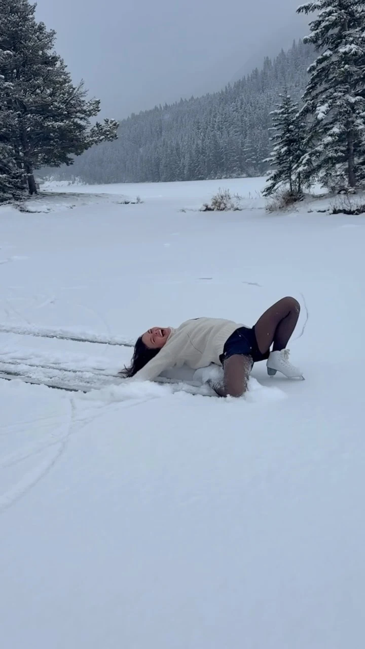 Person lying in snow wearing a cream sweater, dark shorts, sheer tights, and white ice skates.