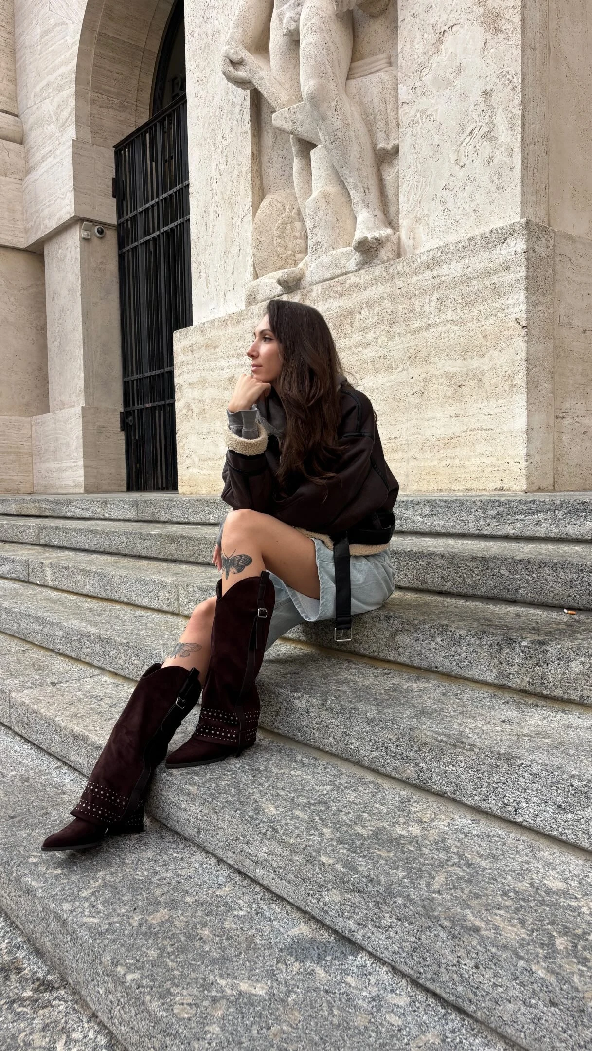 Dark jacket with fuzzy cuffs, pale denim shorts, and tall burgundy studded boots on stone steps.