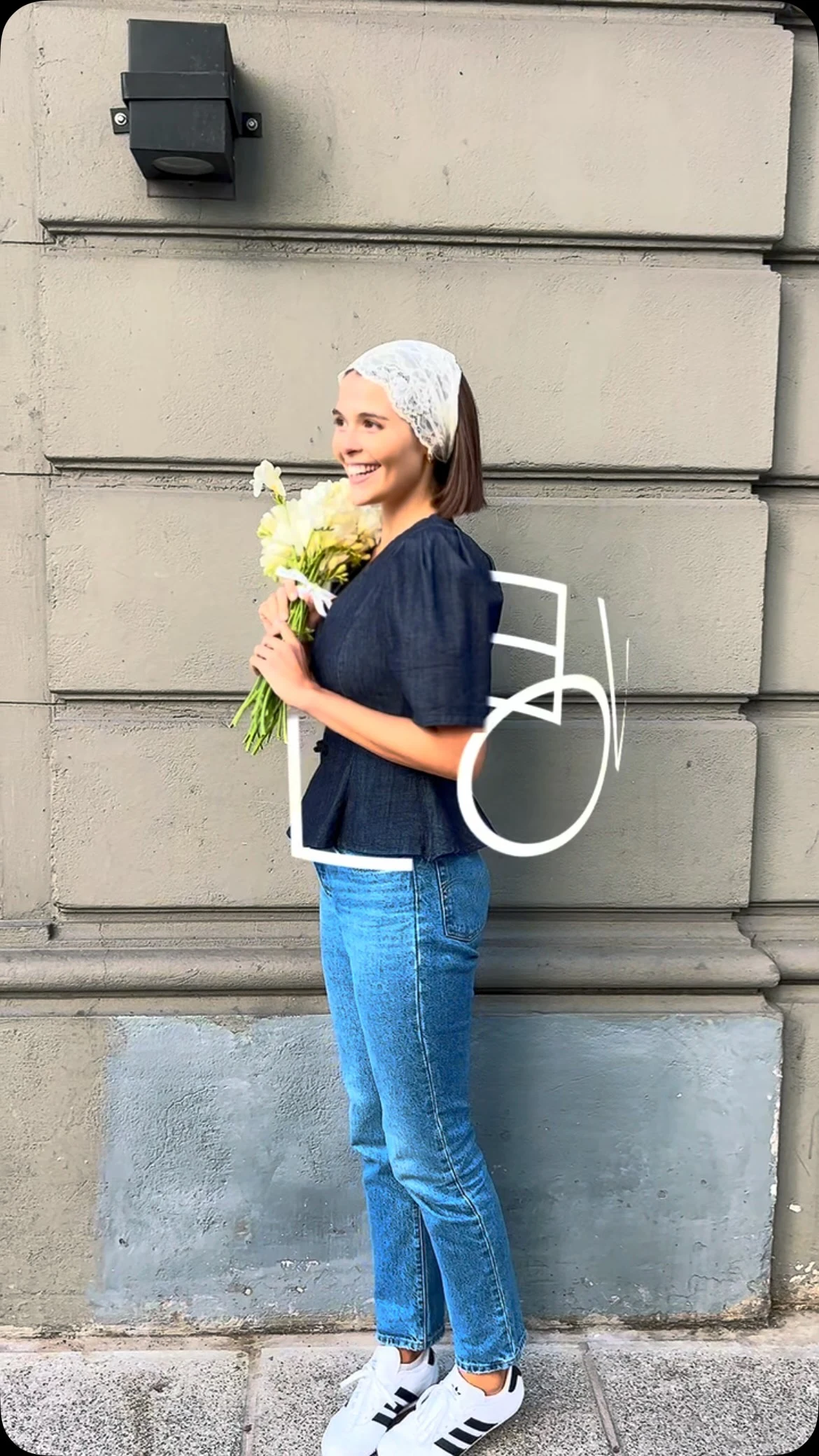 Person in a white lace headscarf, navy blouse, blue jeans, and white sneakers holding pale flowers.