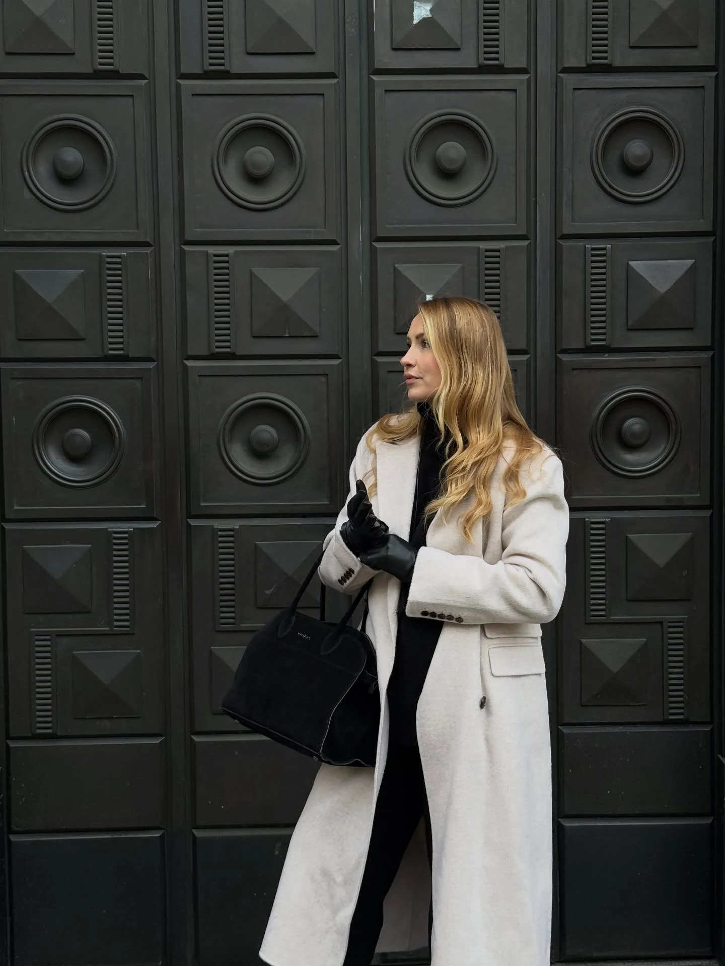 Person in a light long coat over an all-black outfit, standing before a dark paneled door.