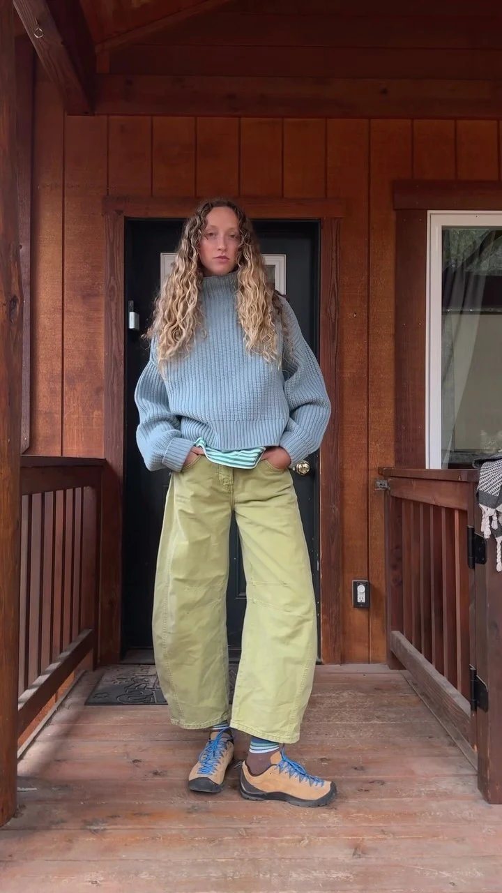 Person on a wooden porch wearing a pale blue chunky sweater, mint top, yellow-green trousers, and blue sneakers.