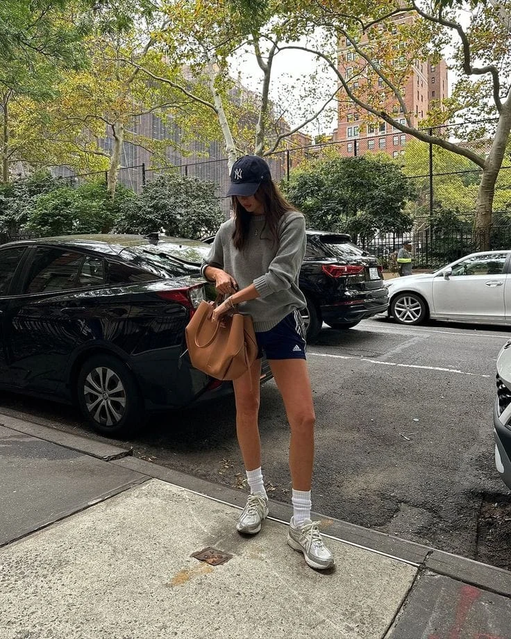 This sporty yet relaxed outfit consists of an oversized grey sweatshirt layered over black Adidas running shorts. The New York Yankees cap, white socks, and silver athletic sneakers complete the effortlessly cool, on-the-go vibe.