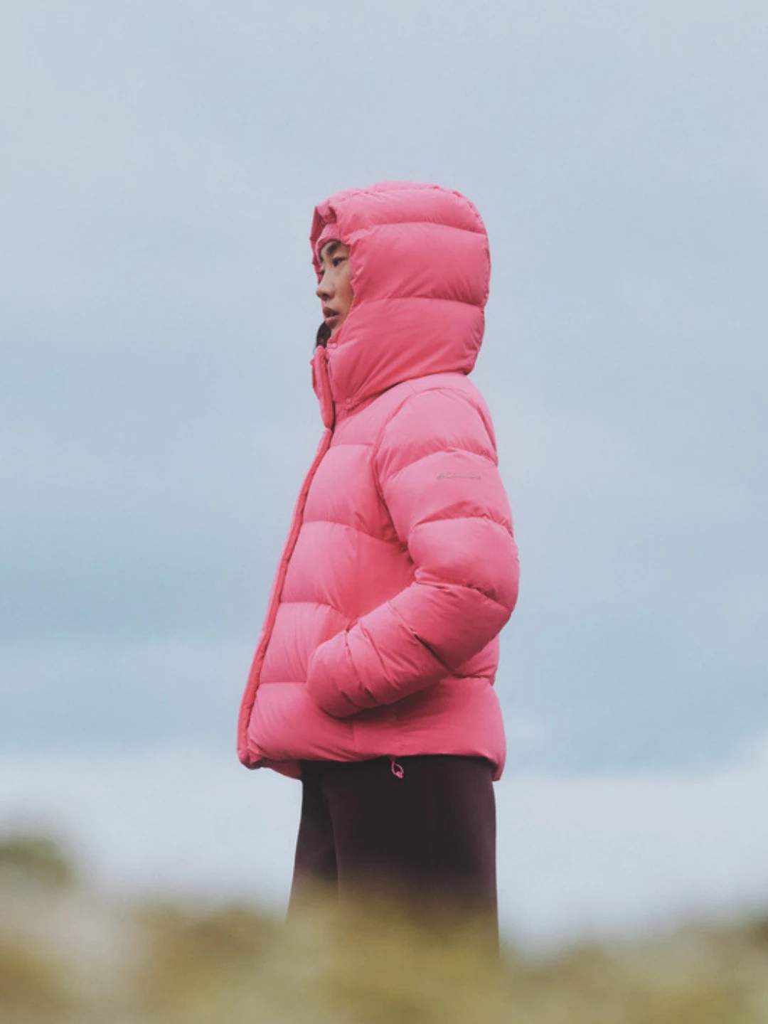 Bright pink hooded puffer jacket with quilted sections worn with dark loose pants outdoors.