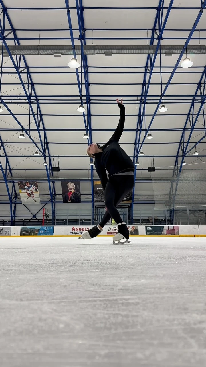 Skater in a fitted black top and leggings with white figure skates posing on an indoor ice rink.