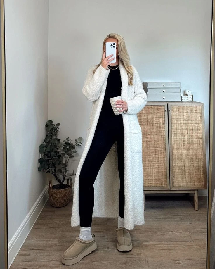 This monochrome outfit features a black fitted top with matching leggings, layered under a long, white fuzzy cardigan for extra coziness. Styled with cream UGG Tasman slippers and white socks, it’s the ultimate lounge-day look.