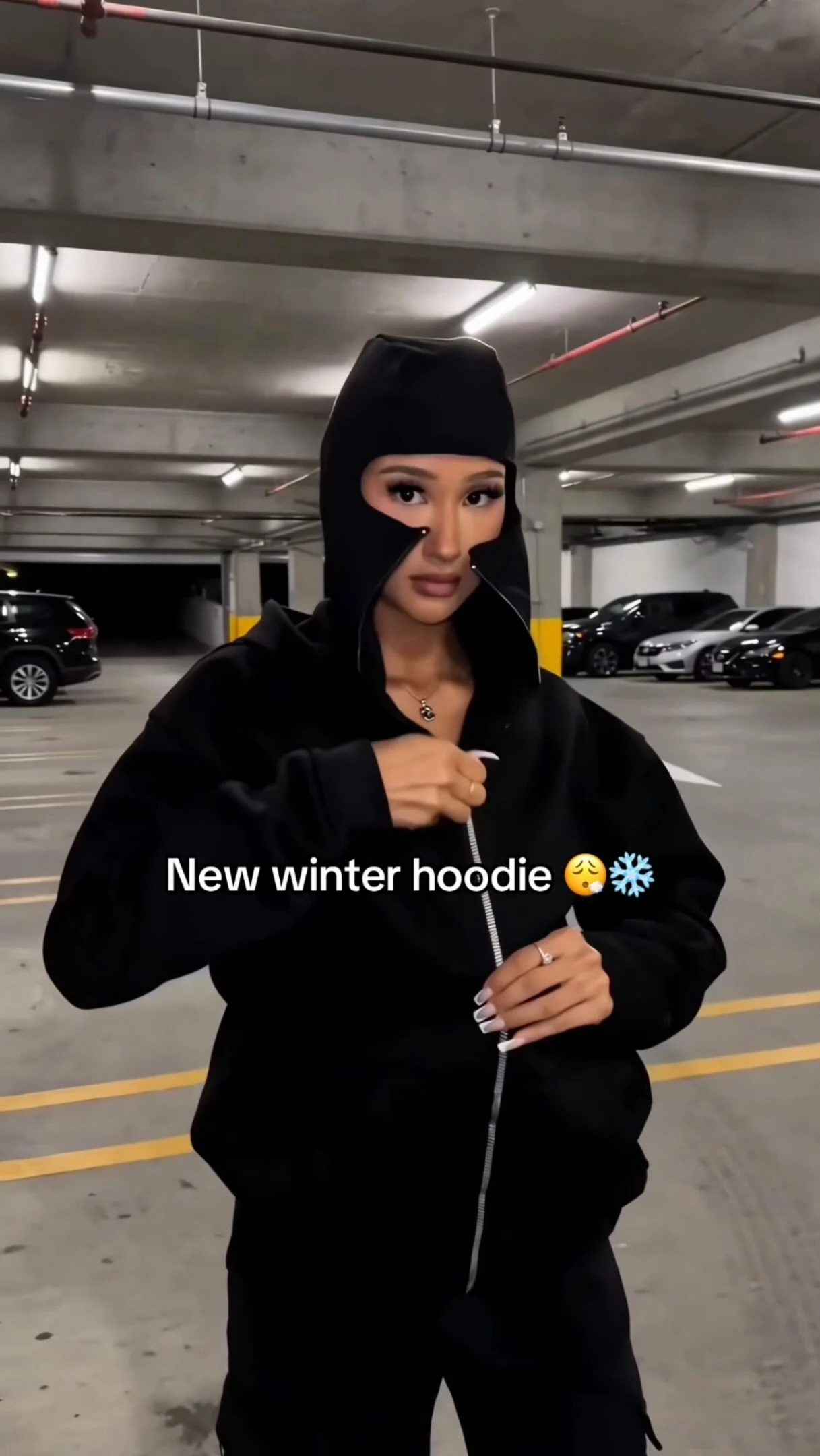 Loose black zip hoodie with face-covering hood worn with matching loose black pants and a pendant necklace.