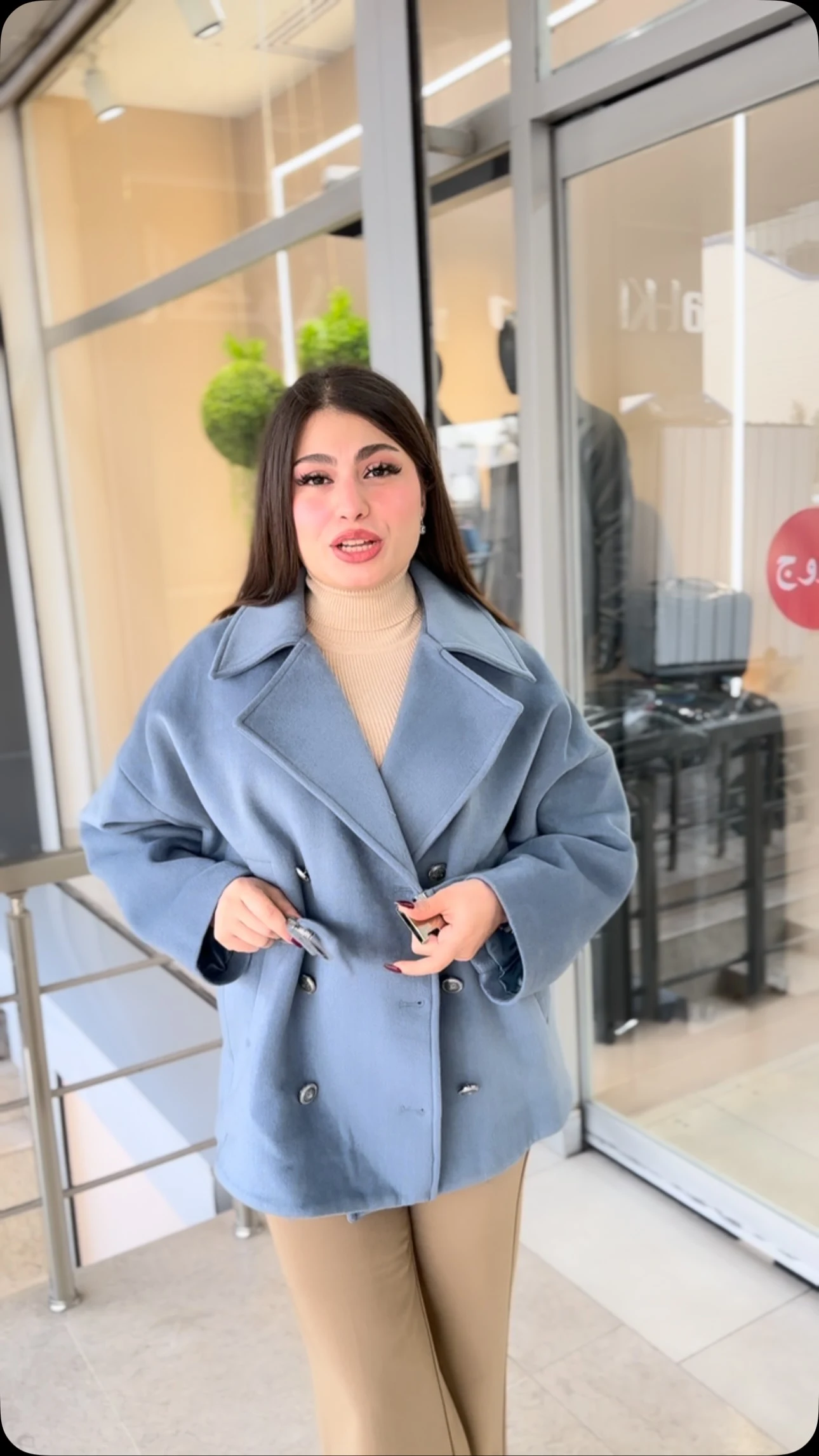 Woman in a powder-blue double-breasted coat over a beige top and slim beige trousers near glass doors.