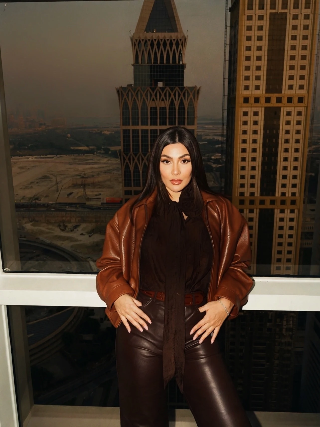 Woman in a brown leather bomber jacket, dark neck-tie blouse, brown leather pants, and brown boots.