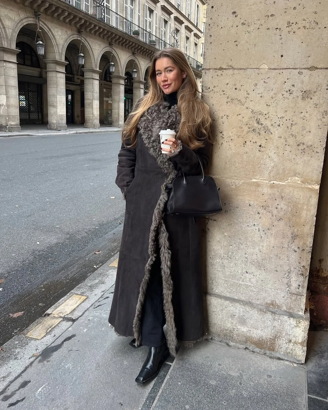 Dark shearling-lined long coat over black trousers and sleek leather boots with a simple black handbag.
