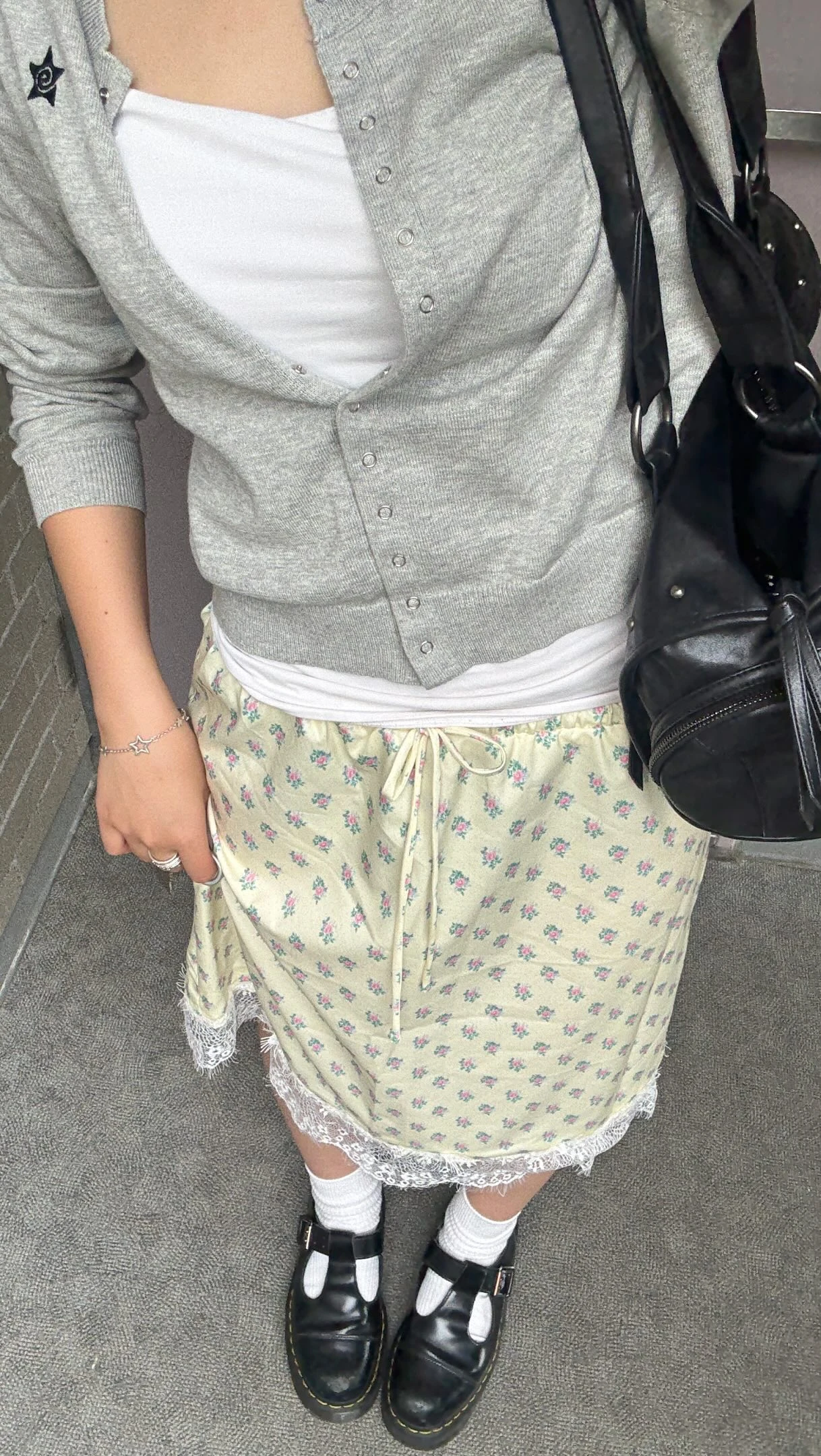 Model wearing a gray cardigan over a pale yellow floral skirt with white socks and black buckled shoes.