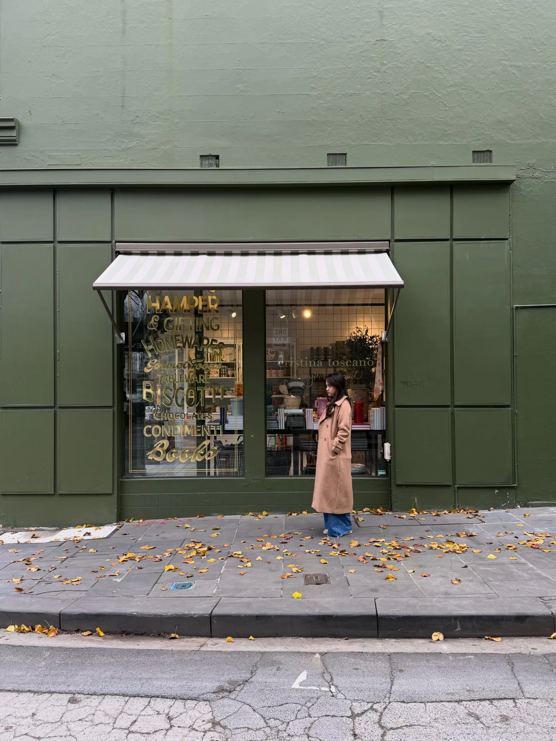 Person in long open camel coat and blue trousers standing outside a storefront with autumn leaves.