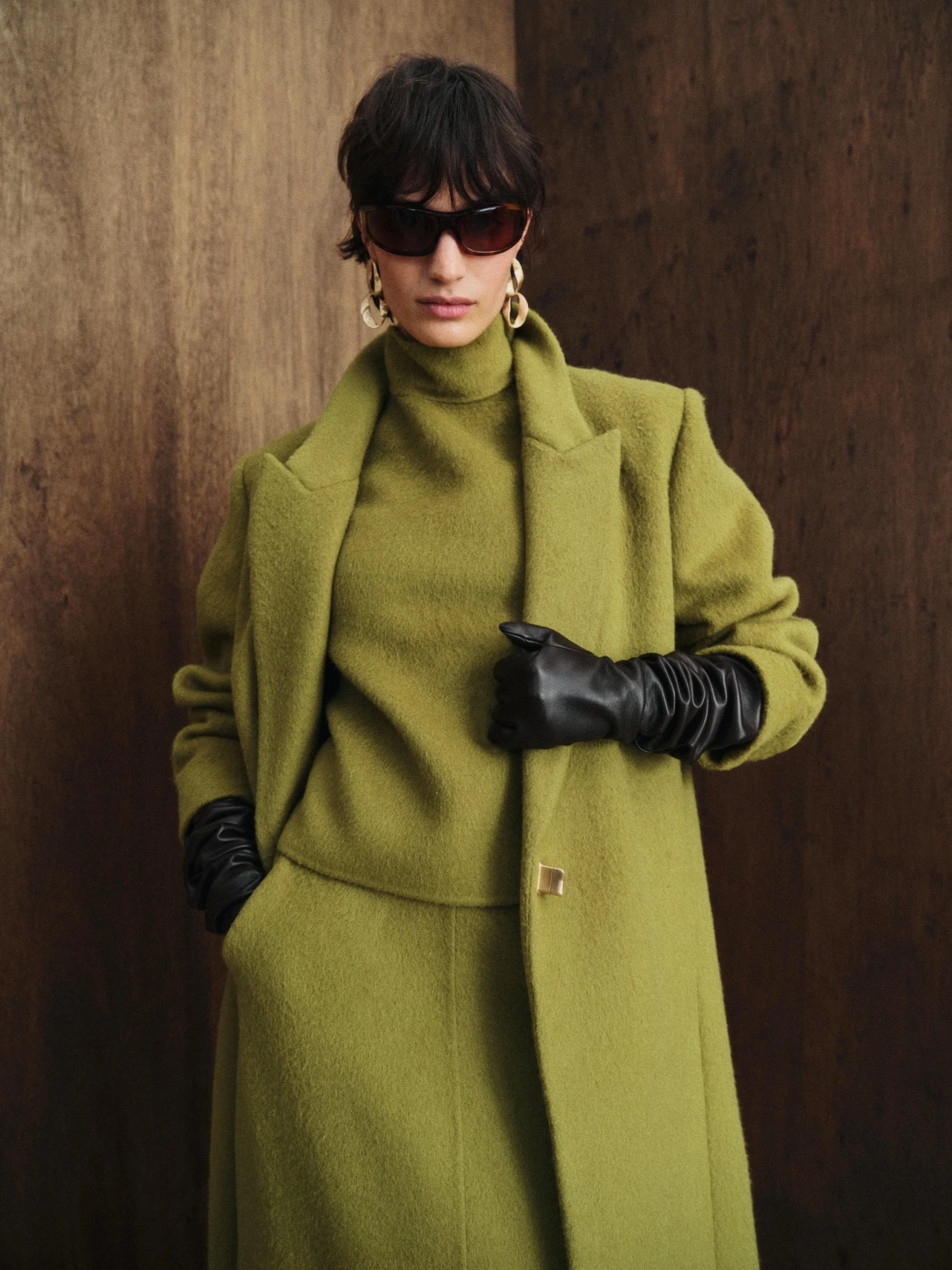 Long olive green coat worn over a matching turtleneck and skirt with black gloves and gold hoops.