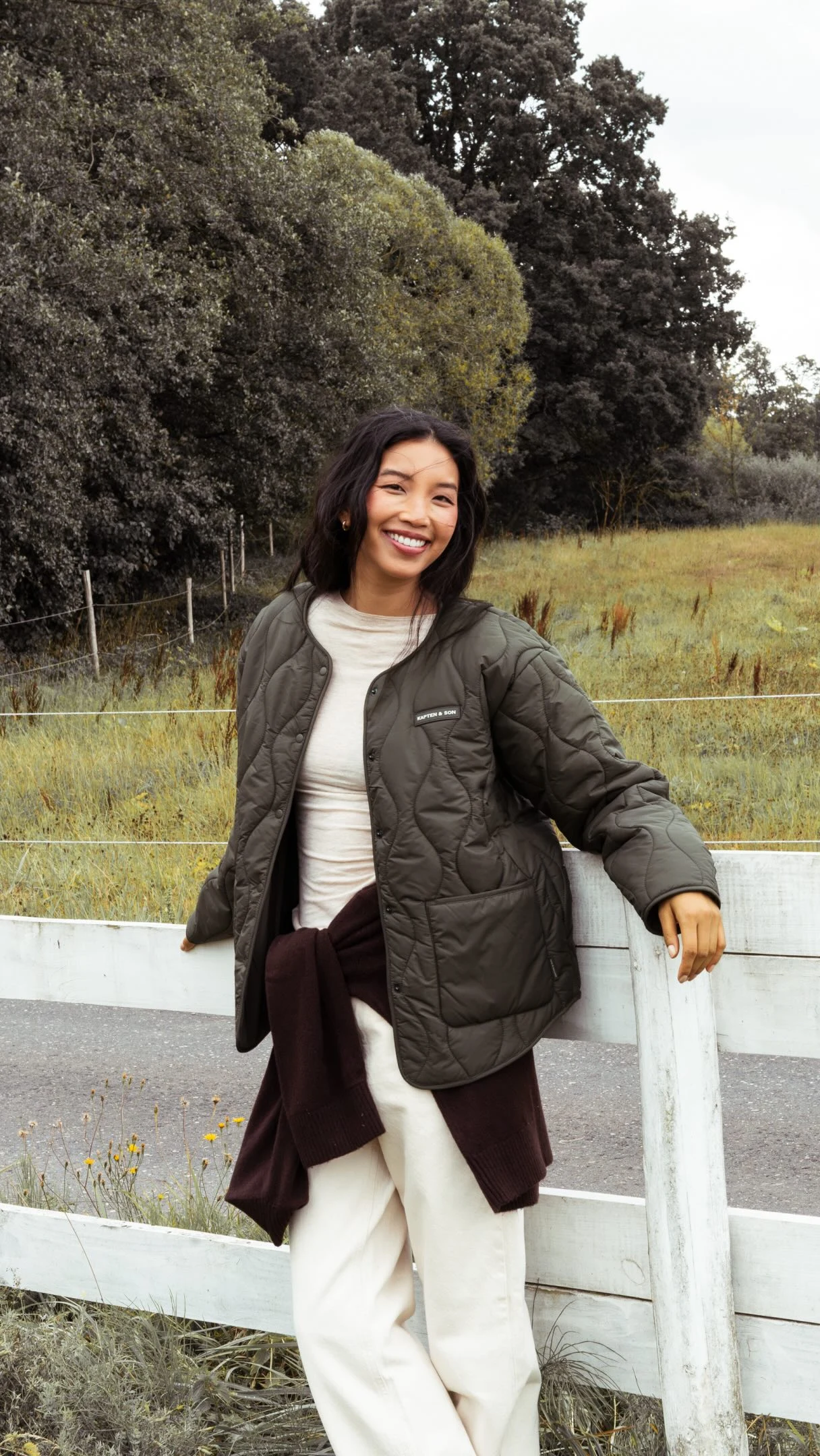 Woman in olive quilted jacket, cream top and trousers, with a burgundy garment tied at her waist.