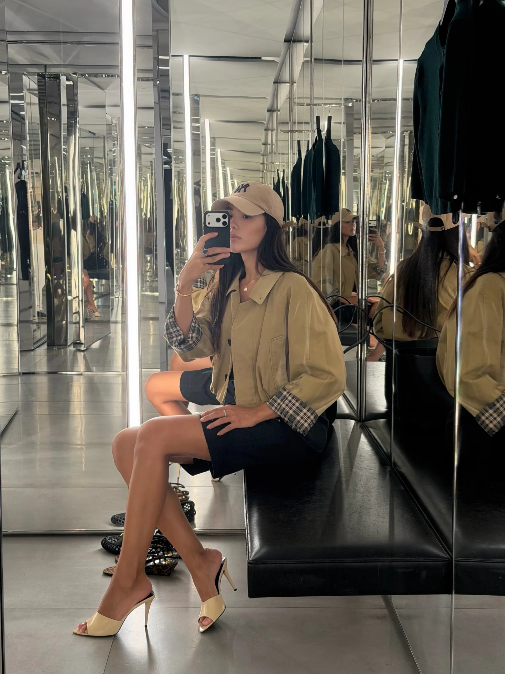 Person in cropped tan jacket, black shorts, beige heels, and tan cap seated on a bench.