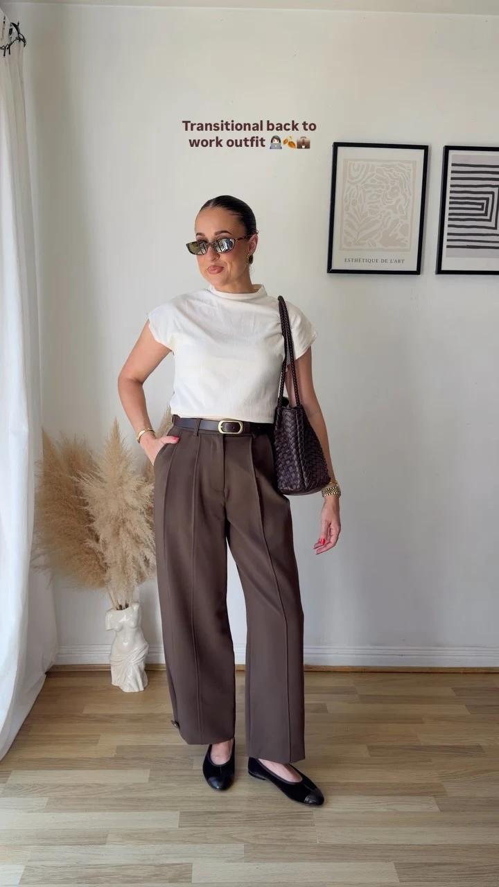 The outfit pairs a loose white short-sleeve T-shirt with high-waisted dark brown trousers featuring front pleats, a belt, and wide straight legs. The trousers fall to the ankles above black flat loafers with a small metallic detail. A dark textured shoulder bag hangs at one side, and the person wears narrow tinted sunglasses and a bracelet. The background includes a pale wood floor, a white curtain, a vase with dried stems, and two framed prints on a light wall.