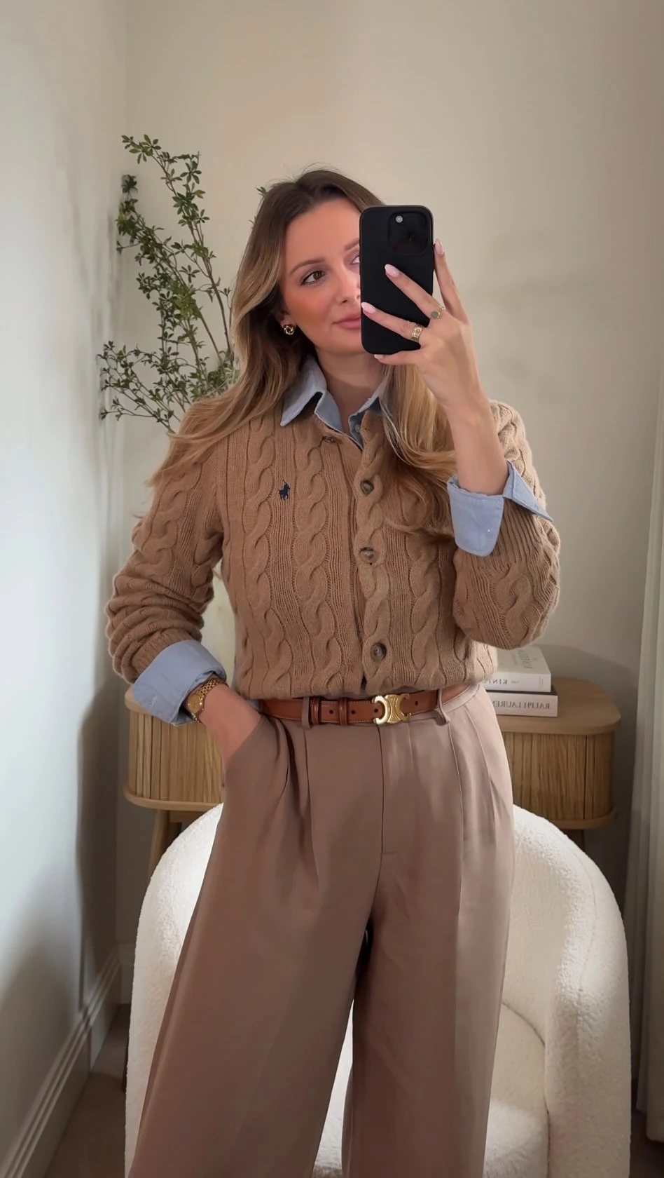 Beige cable-knit sweater over blue collared shirt, tan trousers, brown suede ankle boots, and brown handbag.