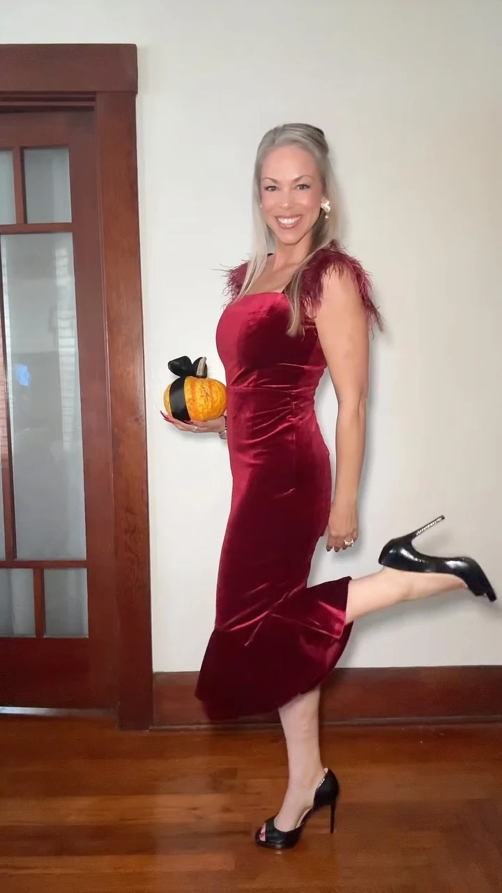 Person in a fitted sleeveless burgundy dress with feathered shoulders, black heels, and an orange pumpkin.