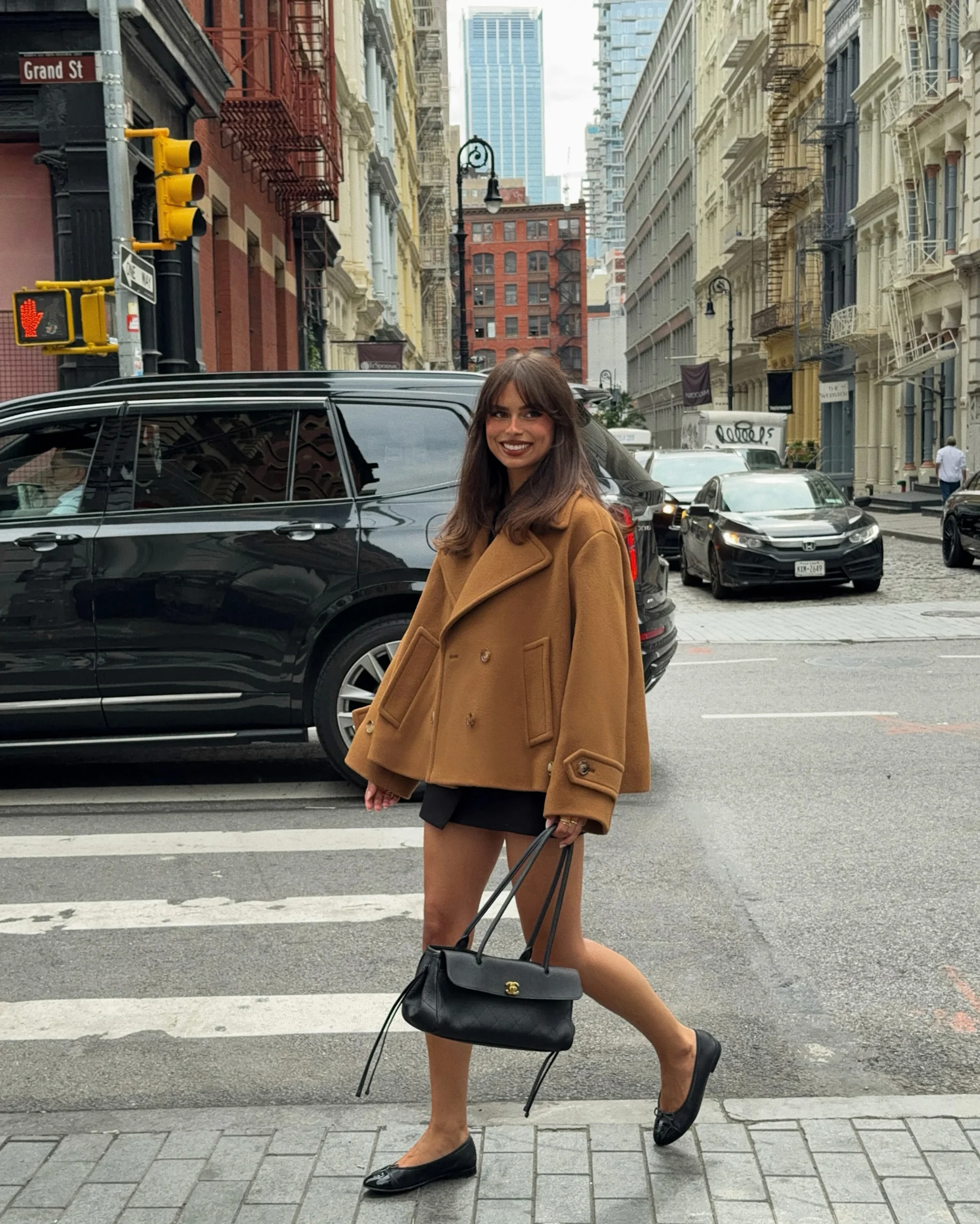 Person in a camel double-breasted coat, black mini skirt, and flat shoes at a city crosswalk.