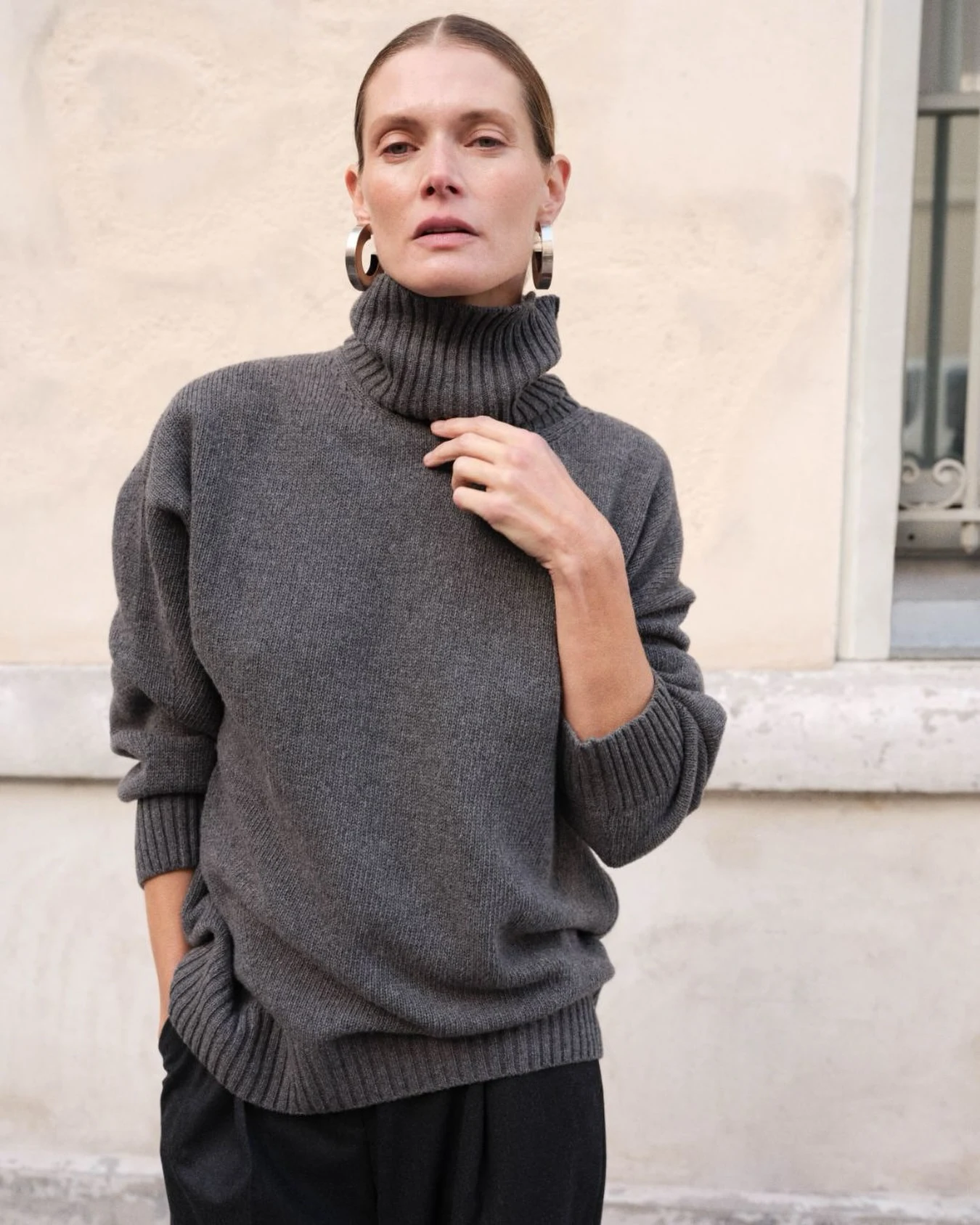 Charcoal oversized turtleneck sweater tucked over loose black trousers with silver hoop earrings.