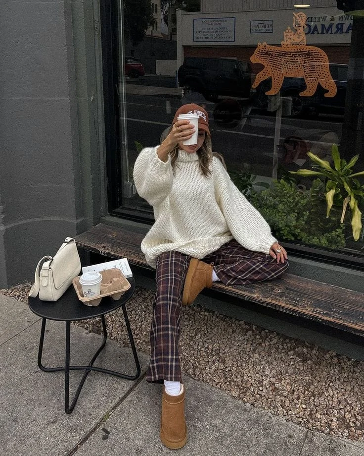 A white oversized chunky knit sweater paired with brown plaid relaxed-fit trousers gives off an effortlessly stylish and cozy vibe. The look is completed with tan UGG boots and a brown baseball cap, making it the perfect relaxed fall outfit.