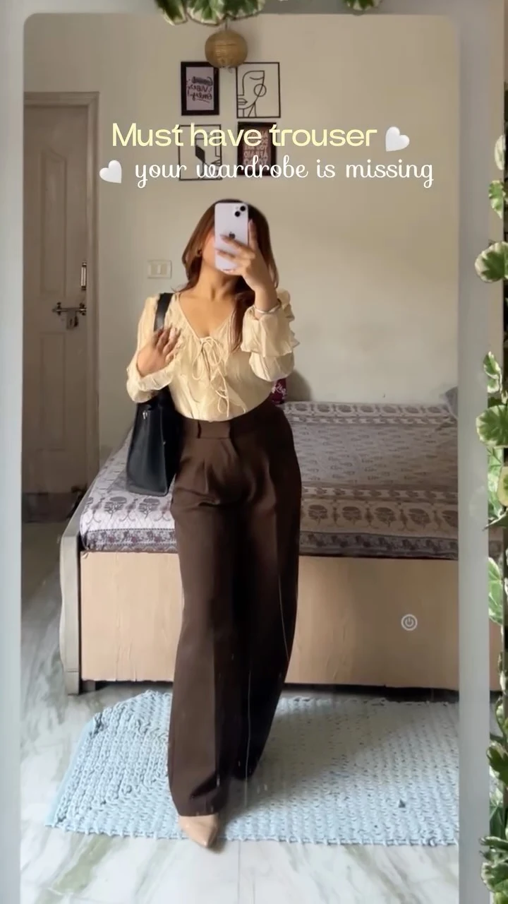 Model in a cream button blouse and dark brown wide-leg trousers with a black shoulder bag.