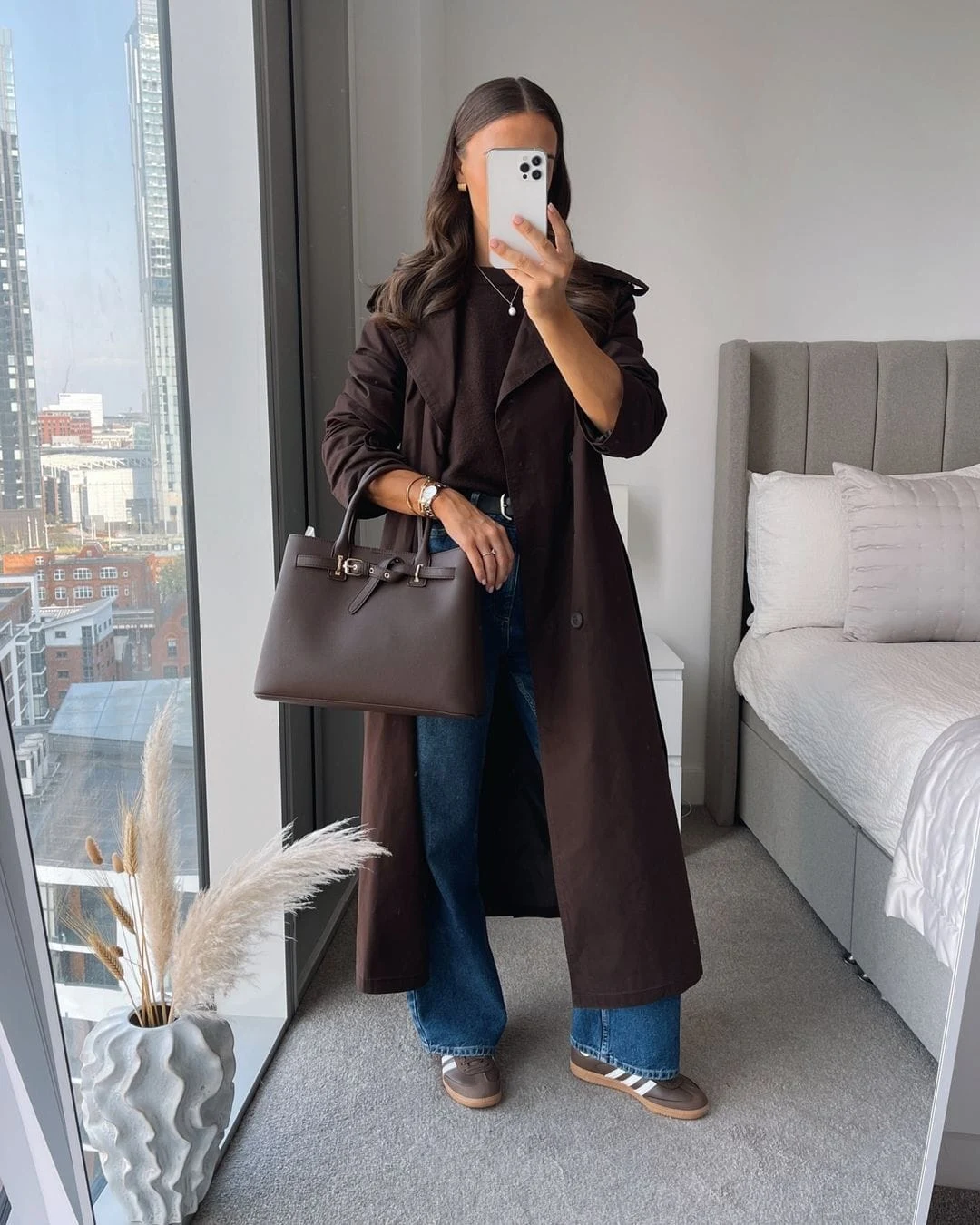 A dark brown trench coat layered over casual jeans and sneakers makes this outfit a blend of comfort and style. The brown leather handbag adds a touch of elegance to this laid-back winter look.