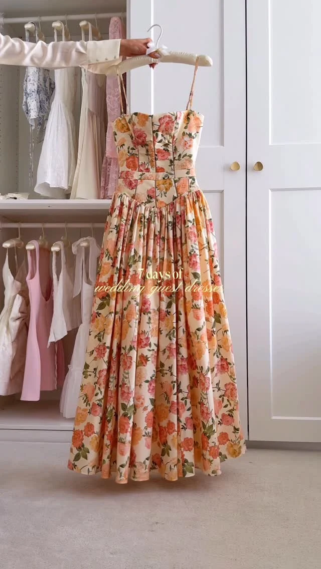 A full-length strapless dress hangs on a white hanger in front of a wardrobe. Its fitted bodice has visible vertical seams, and the gathered skirt falls in broad pleats. The cream-colored dress is covered in a large floral print with coral, peach, pink, and green tones.