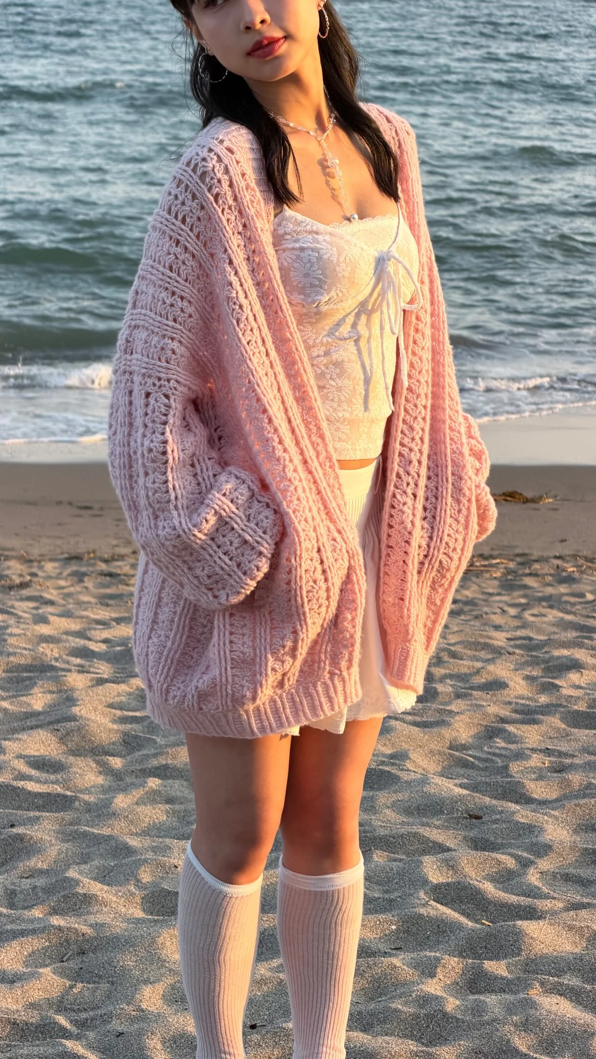 Oversized pink crochet cardigan over a lace top, short skirt, and knee-high socks on a beach.