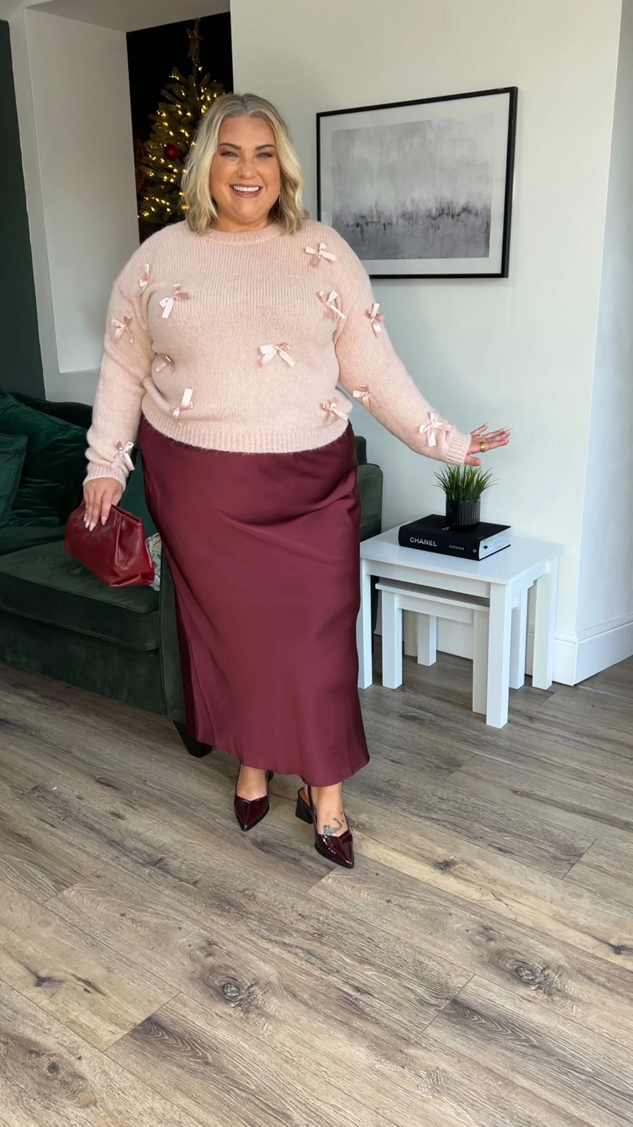 Person wearing a pink bow sweater with a burgundy midi skirt and pointed-toe shoes near a Christmas tree.
