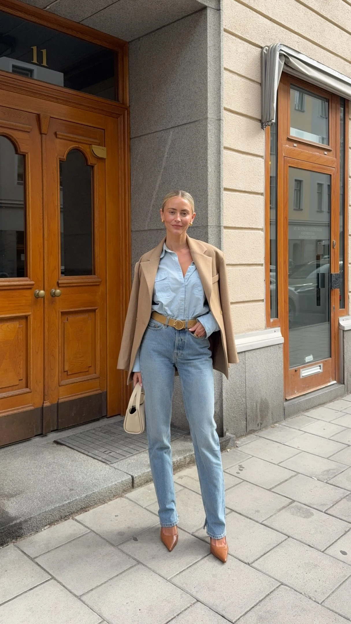 Light blue shirt tucked into pale jeans, beige coat open, brown pointed shoes, cream bag.
