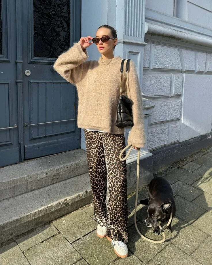 This outfit pairs a beige oversized knit sweater with bold leopard-print wide-leg pants for a cozy yet stylish statement. Accessories like white sneakers, black sunglasses, and a quilted shoulder bag add a trendy, relaxed vibe.
