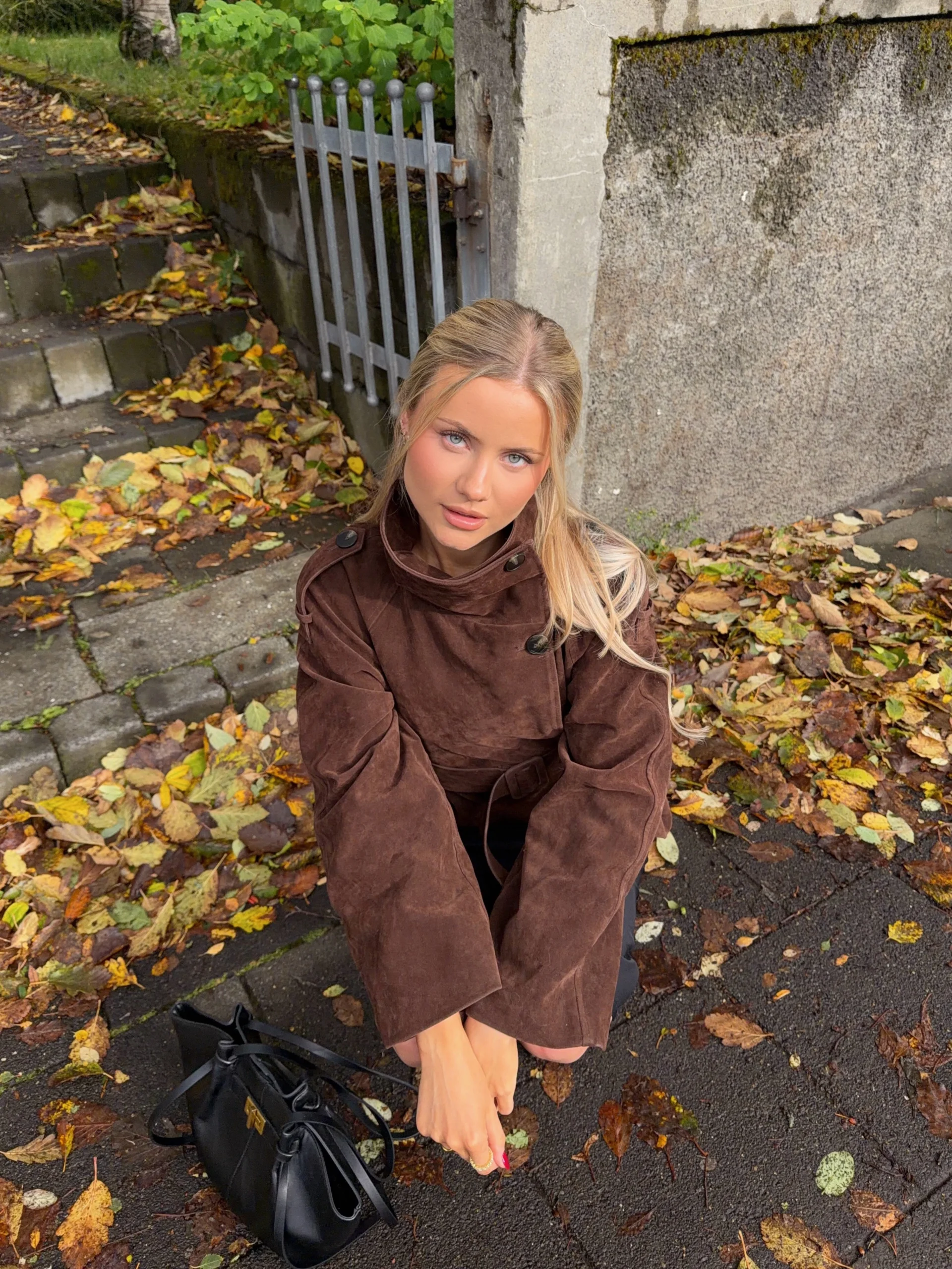 Dark brown belted coat with wide sleeves and folded collar, black handbag on leaf-covered pavement nearby.