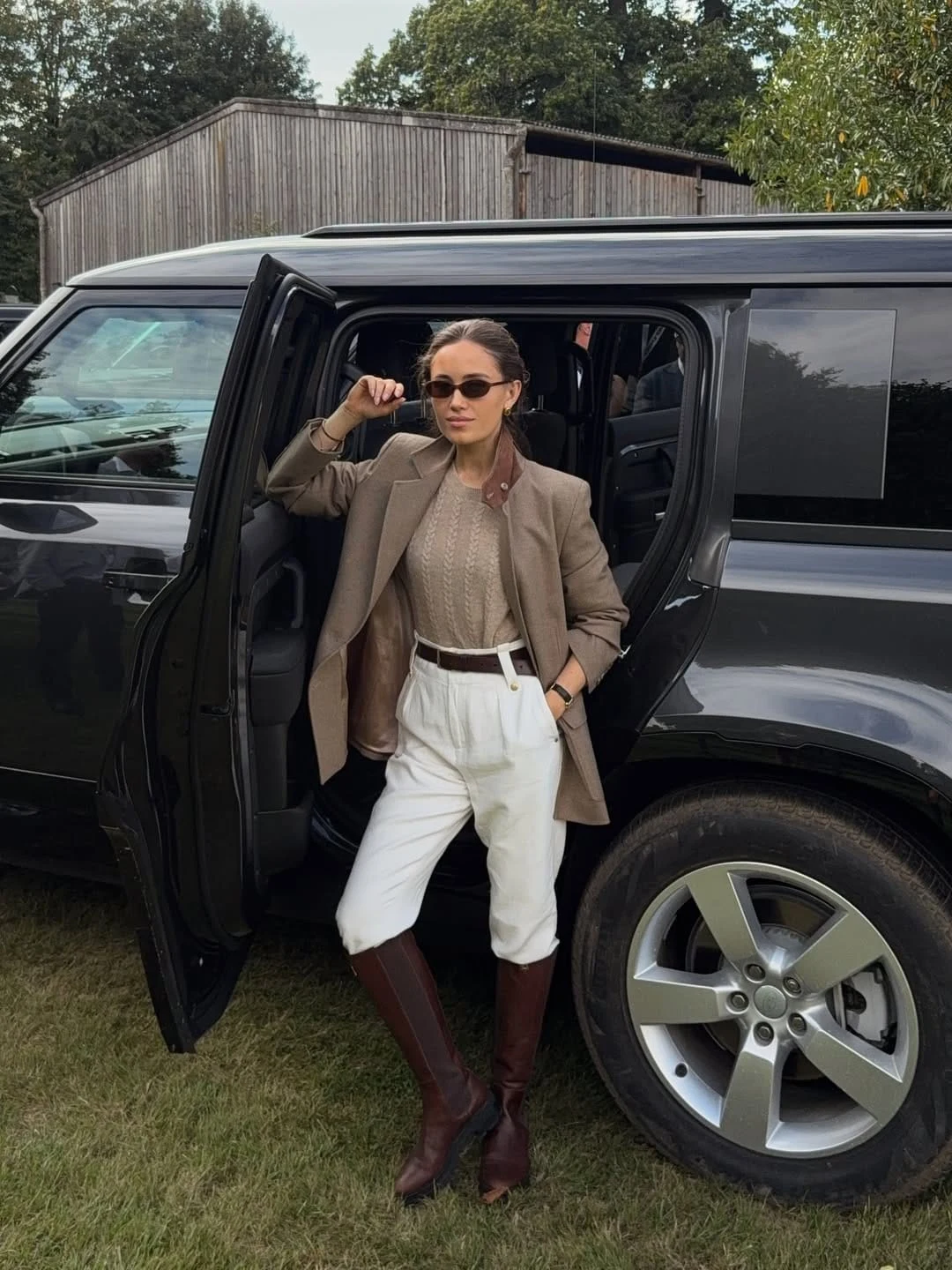 A cable-textured knit top layered under a structured neutral blazer, with relaxed white trousers and tall brown riding boots, is the combination that works best when you want country chic to read as polished and slightly formal rather than weekend-relaxed — the blazer over the knit creates a layered formality, and the white trousers lighten the whole palette so it feels bright and considered rather than heavy. The textural contrast between the knit and the smooth blazer fabric is subtle but significant, adding a visual dimension that a plain shirt underneath the same blazer would not provide. I love this with oversized sunglasses for that quietly confident countryside-with-attitude energy.