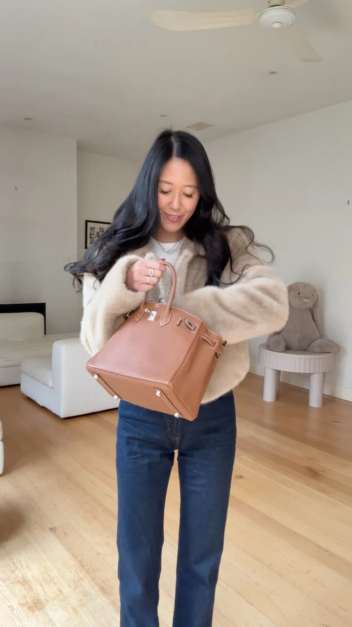 Loose cream cardigan worn open over a light top and straight-leg dark blue jeans, tan handbag held at front.