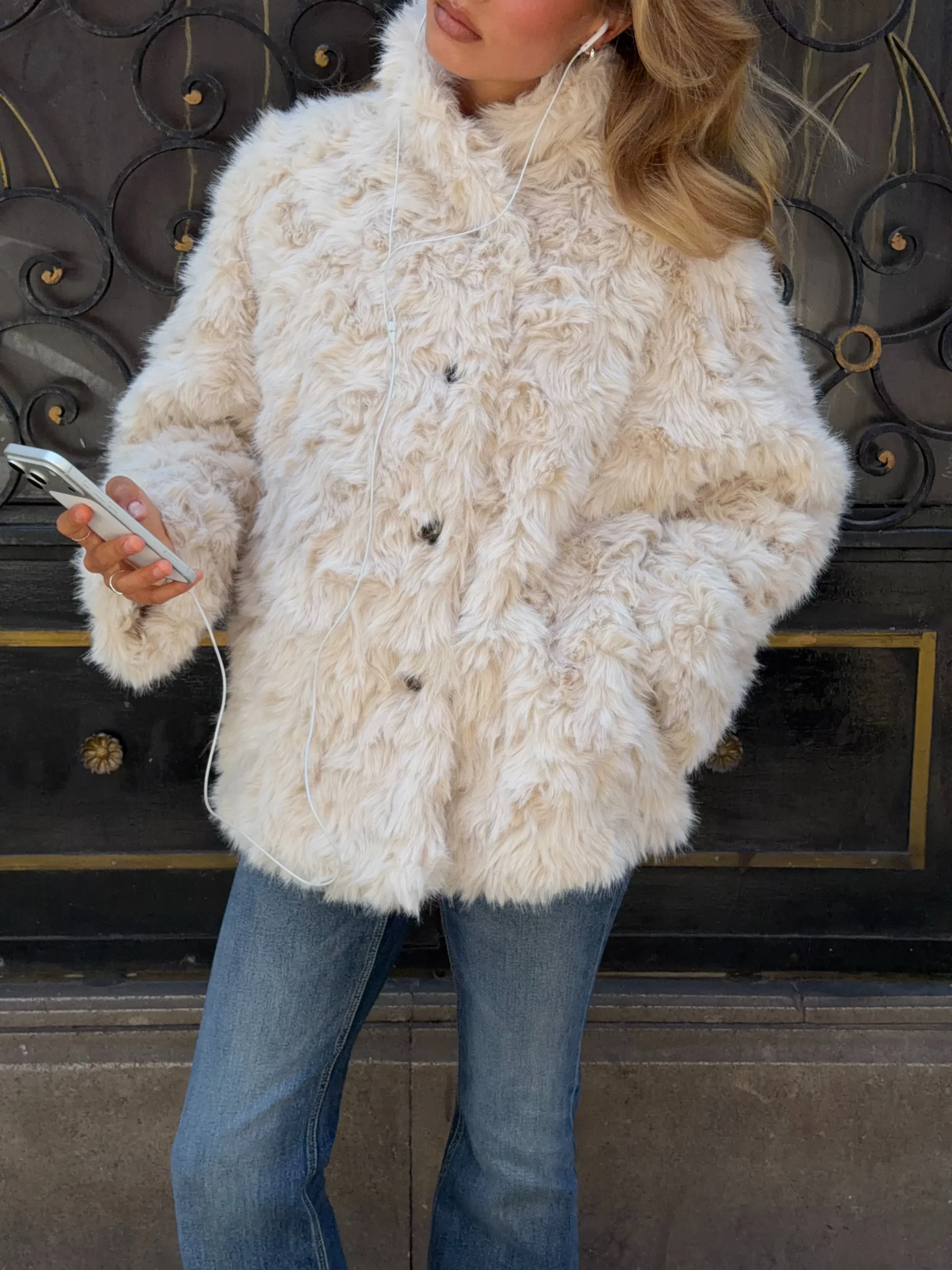 Woman in a cream shaggy faux-fur jacket, white tee, light-wash flared jeans, and white sneakers.