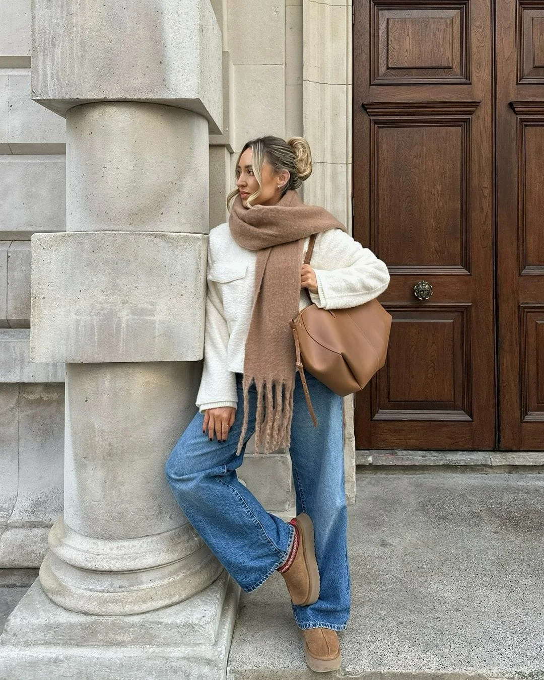 This cozy look includes a cream sweater paired with light blue jeans and tan boots. The oversized beige scarf and matching tan handbag complete the casual yet polished winter style.