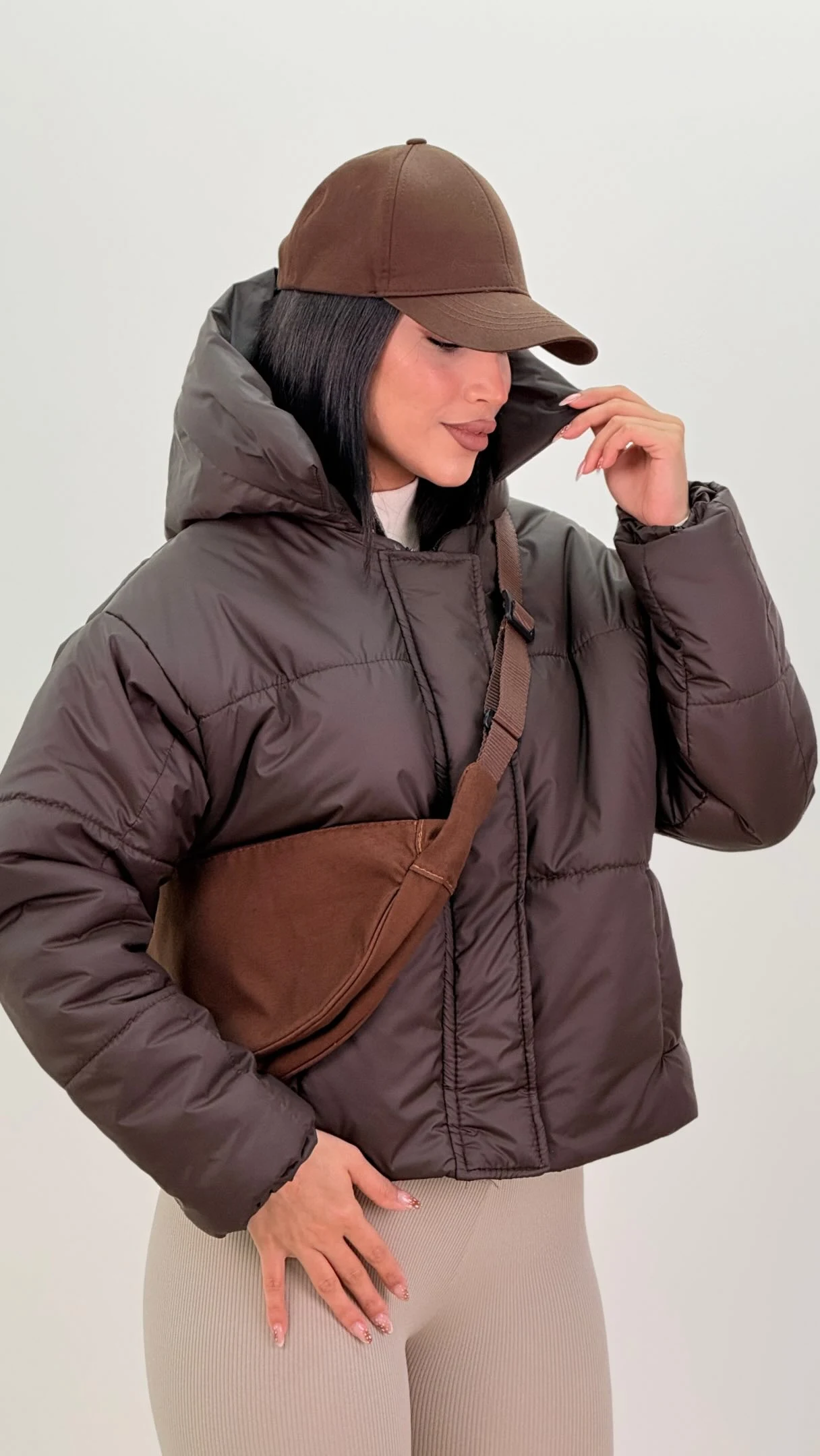 Dark brown hooded puffer jacket styled with beige ribbed leggings, brown crossbody bag, and matching baseball cap.