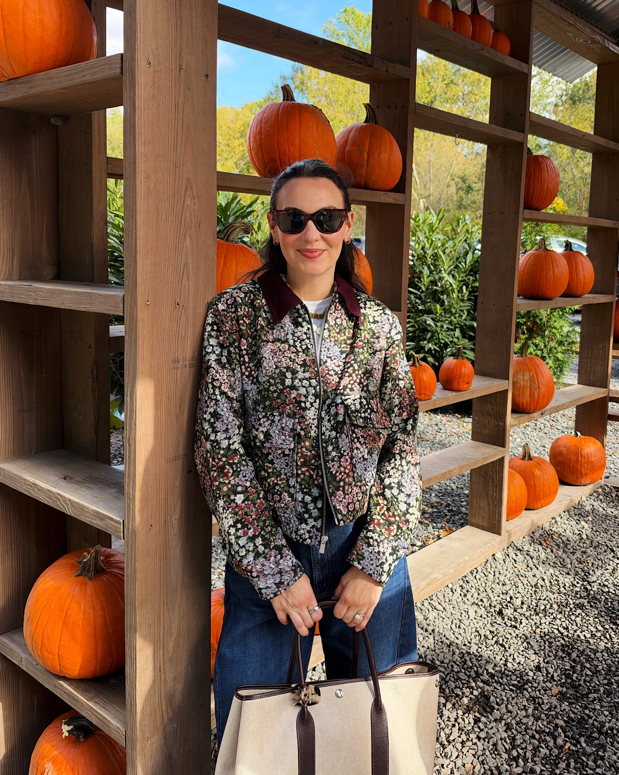Woman in a floral zip-up jacket with maroon collar, white tee, blue jeans, and a canvas tote.