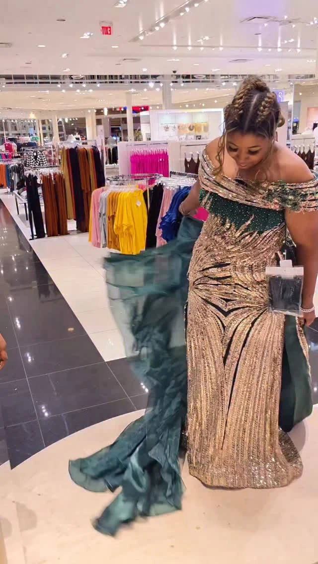 Person wearing a fitted dark green off-shoulder gown with gold geometric embellishment and a sheer train.