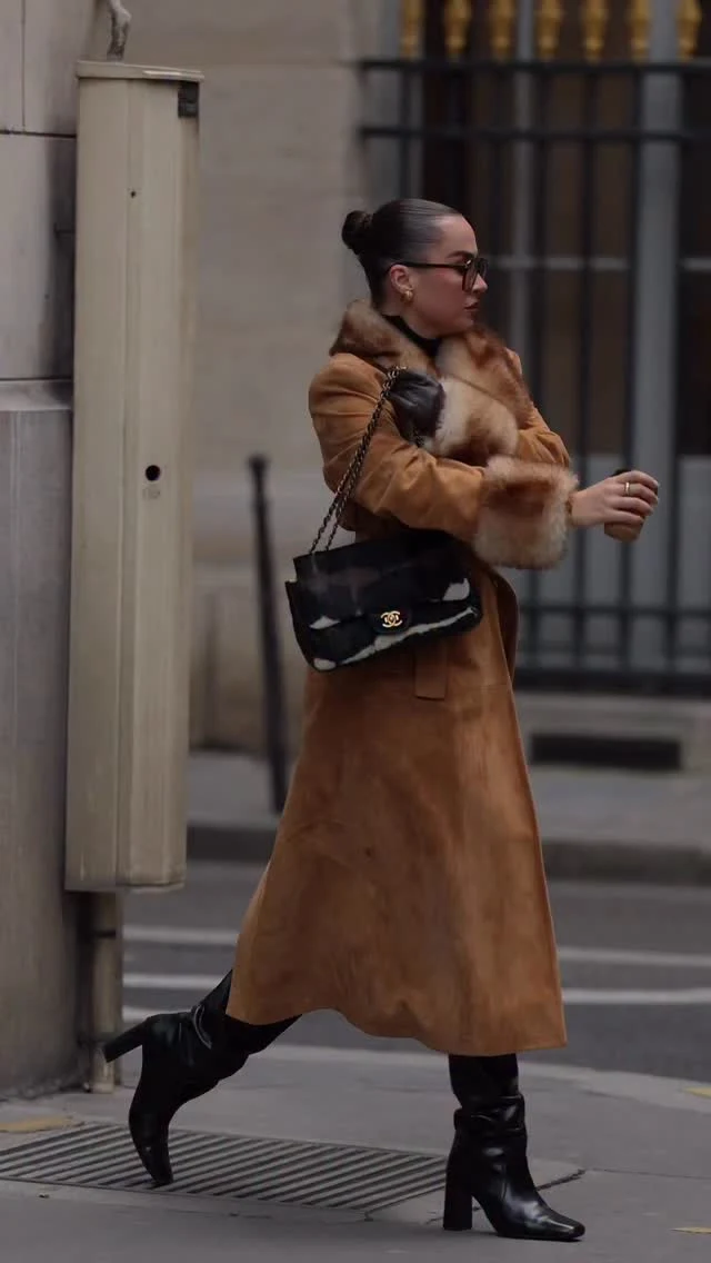 Person wearing a long tan coat with plush collar, black knee-high boots, and a chain-strap bag.