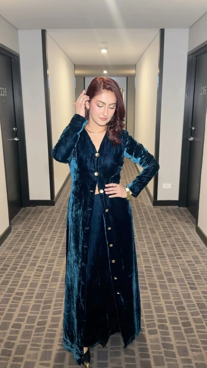 A long, deep teal dress has a plush-looking surface, long sleeves, a V-shaped neckline, and a row of gold-tone buttons down the front. The buttoned front opens slightly near the hem, revealing dark pointed shoes. A slim necklace and a gold-tone watch are also visible.