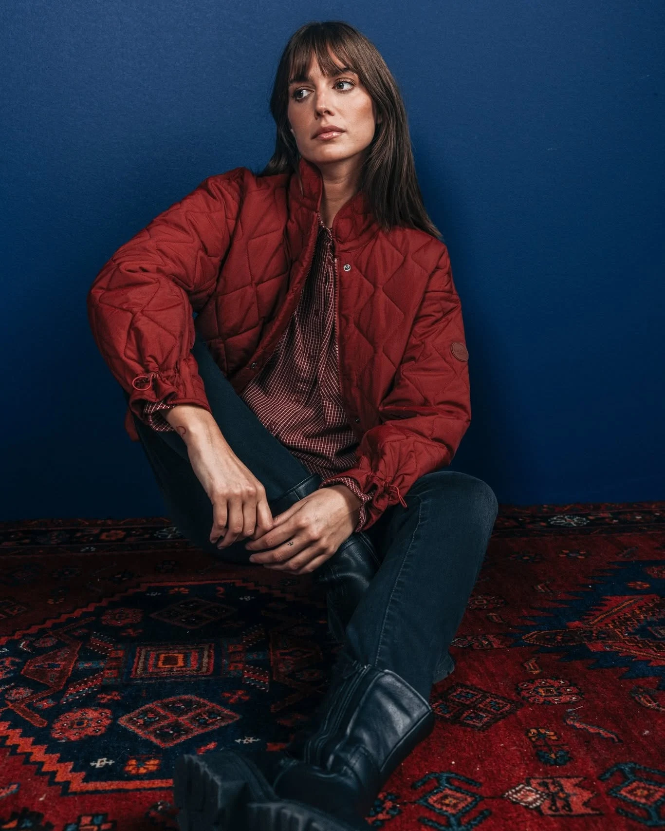 Seated person in a red quilted jacket, striped top, dark jeans, and black boots on a patterned rug.
