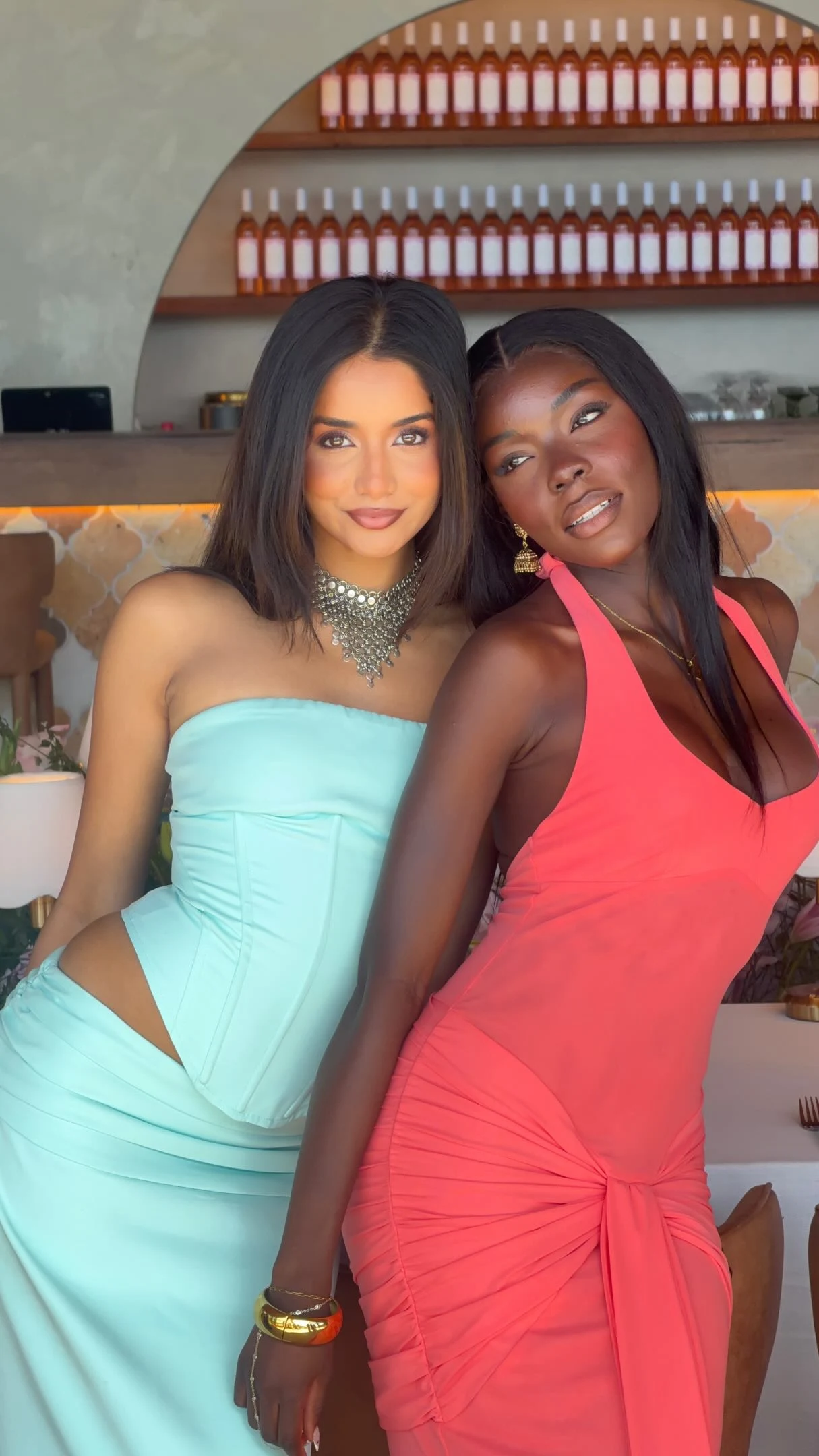 Two women in a bar, one in aqua strapless corset set, one in coral halter dress.