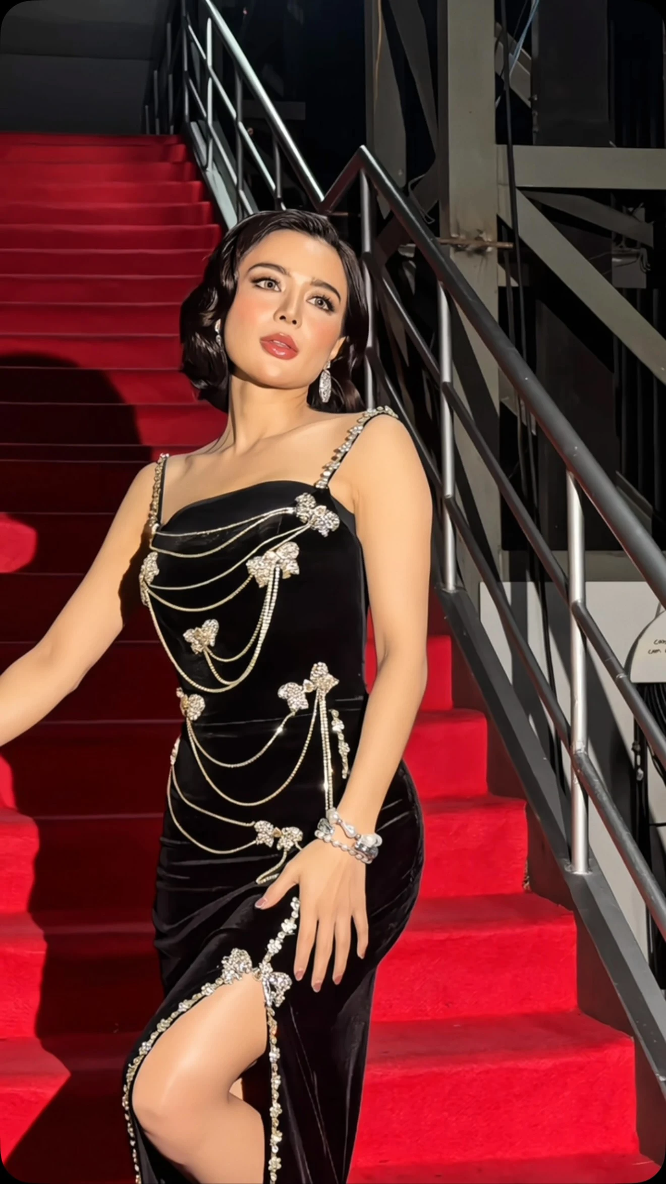 A fitted black gown has thin embellished shoulder straps, a gently curved neckline, and a high slit. Silver-toned chains drape across the bodice and skirt, accented with sparkling floral ornaments. The wearer also has dangling earrings and stacked bracelets. She stands on a red-carpeted staircase with dark metal railings.