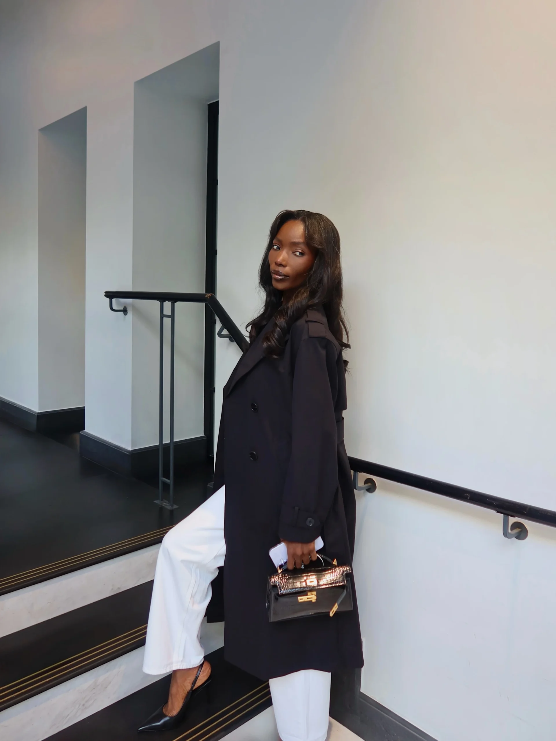 Long black coat with broad lapels over white trousers, black pointed-toe shoes, and small structured black handbag on stairs.