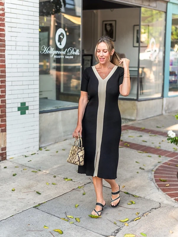 Woman in a black V-neck dress with a beige center stripe, carrying a patterned bag outdoors.