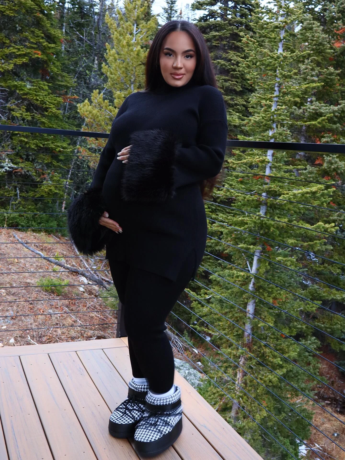 The outfit features a long black sweater with oversized fuzzy cuffs and black fitted pants. Black-and-white patterned boots have thick black soles and folded cuffs. The person stands outdoors on a wooden deck.