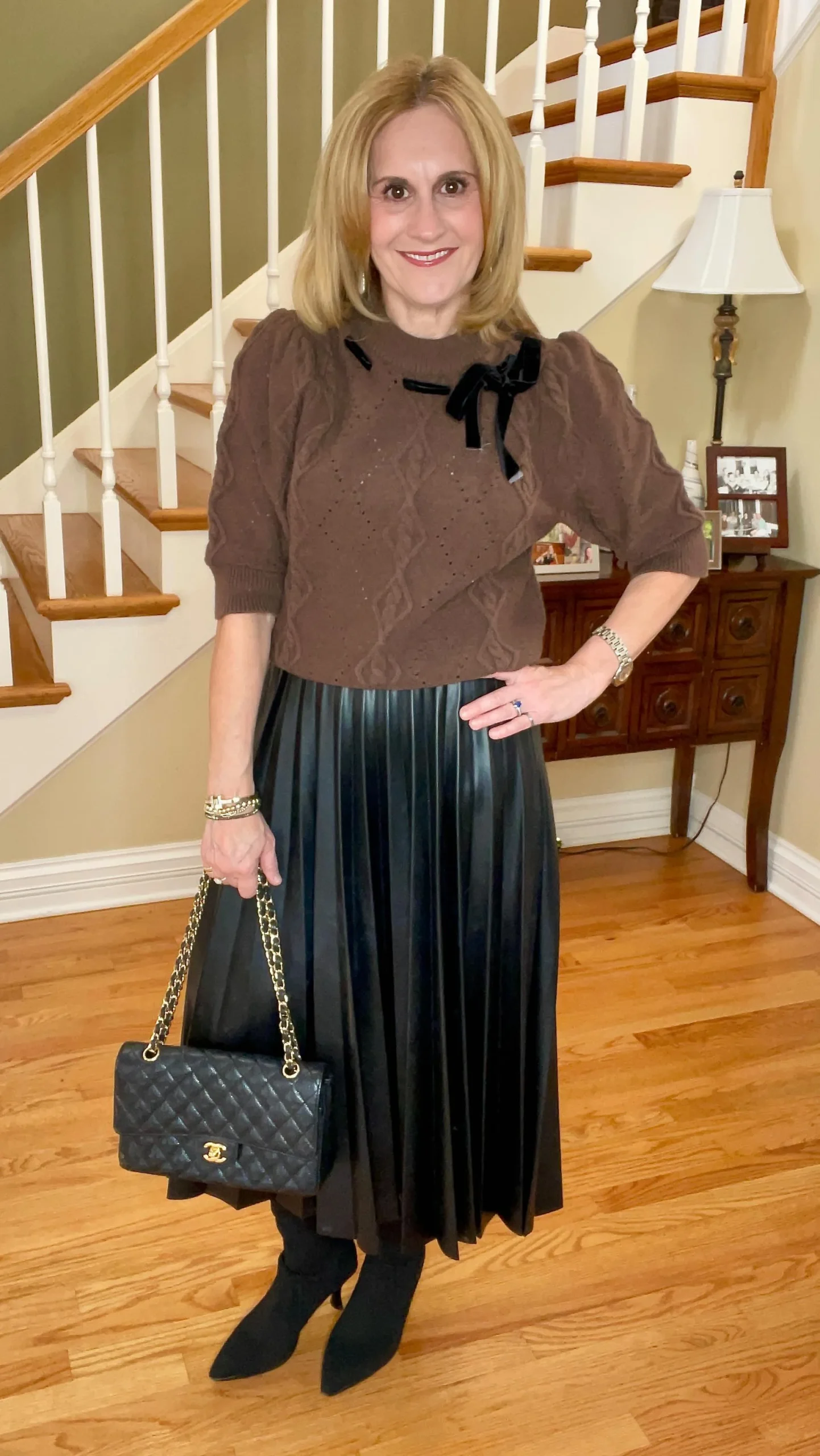 Woman wearing a brown cable-knit sweater with a velvet bow, black faux-leather pleated midi skirt, and ankle boots.